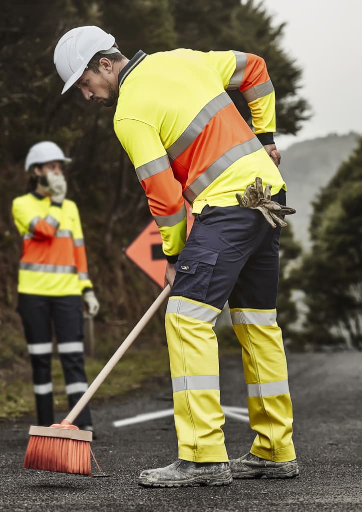 High-visibility yellow and orange work pants with reflective tape, worn by a man sweeping the road, alongside a woman in similar attire.