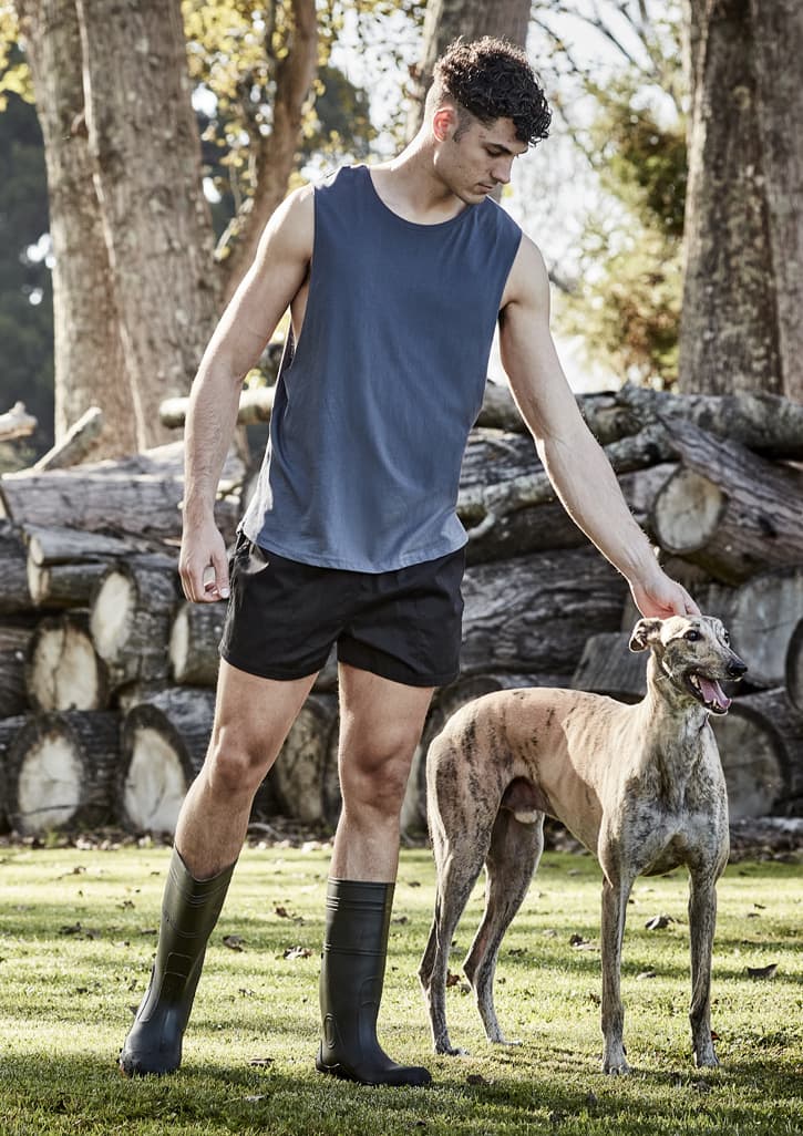 A man in a grey tank top and black shorts interacts with a greyhound in a natural outdoor setting.