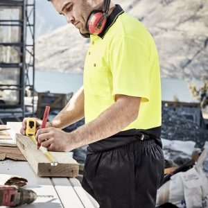A man wearing a high-visibility yellow polo shirt and black rugby shorts measures wood on a construction site.