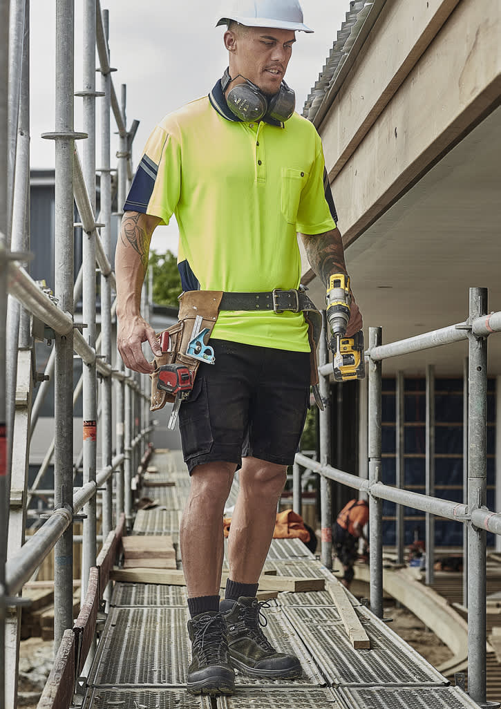 A man wearing a high-visibility yellow polo shirt and black cargo shorts, carrying tools on a construction site.