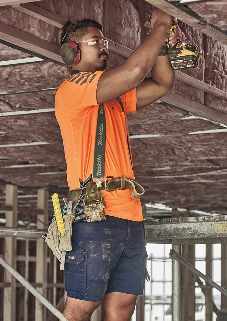 A man wearing an orange shirt and navy blue cargo shorts works on a construction site, using a power drill.