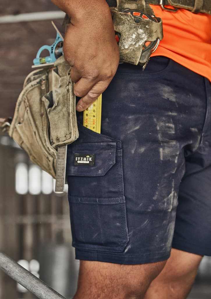 Men's navy cargo shorts featuring multiple pockets, worn with a tool belt, showing a measuring tape and work environment dust.