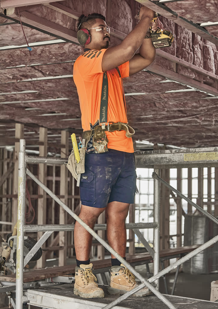 A worker in an orange shirt and navy cargo shorts uses a power tool on a construction site.