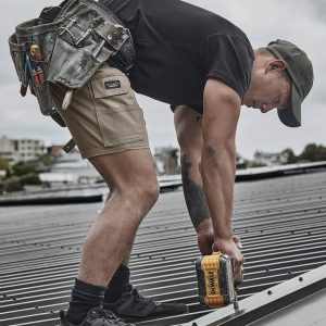 A person wearing khaki cargo shorts and a black t-shirt is using a power drill on a roof. Tools hang from a tool belt.
