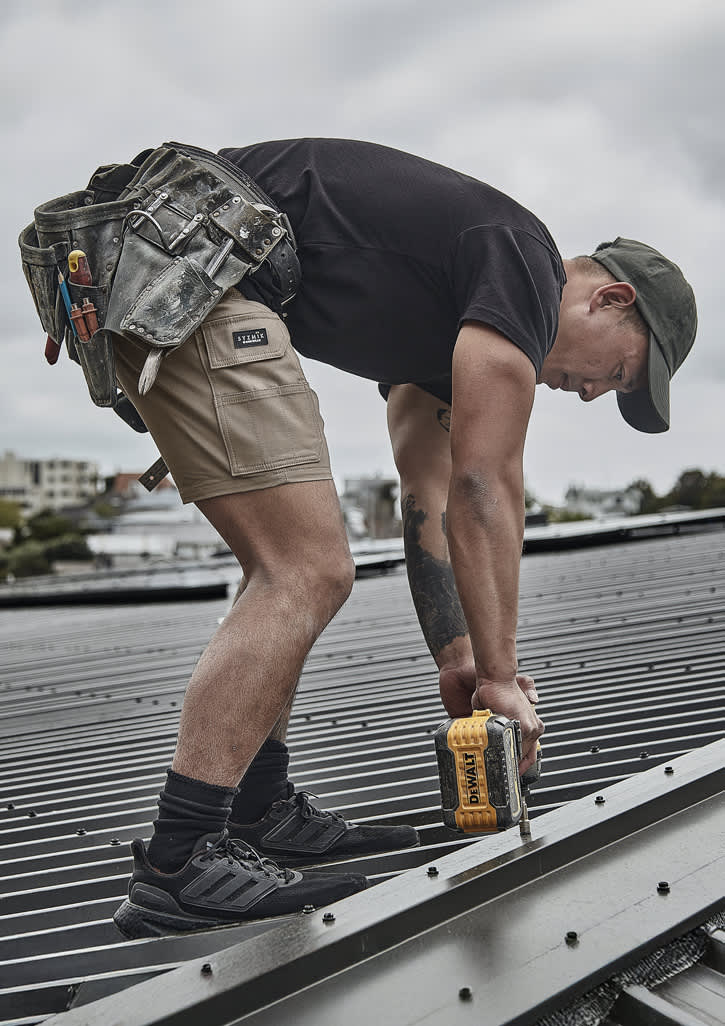 A person wearing khaki cargo shorts and a black t-shirt is using a power drill on a roof. Tools hang from a tool belt.