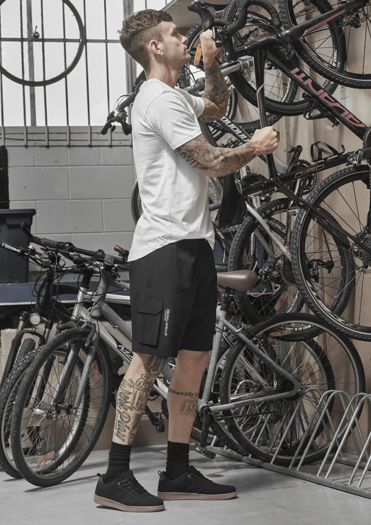 A man stands in a garage adjusting a bike, wearing black Men's Streetworx board shorts and a white t-shirt.