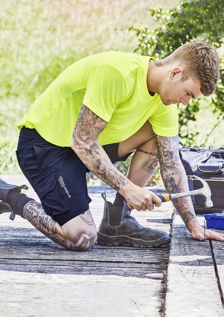 A person kneeling outdoors in bright yellow clothing and dark blue sports shorts, using a saw on a wooden surface.