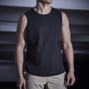 A man wearing a black sleeveless top and beige sports shorts stands in a dimly lit setting. The shorts feature a logo.