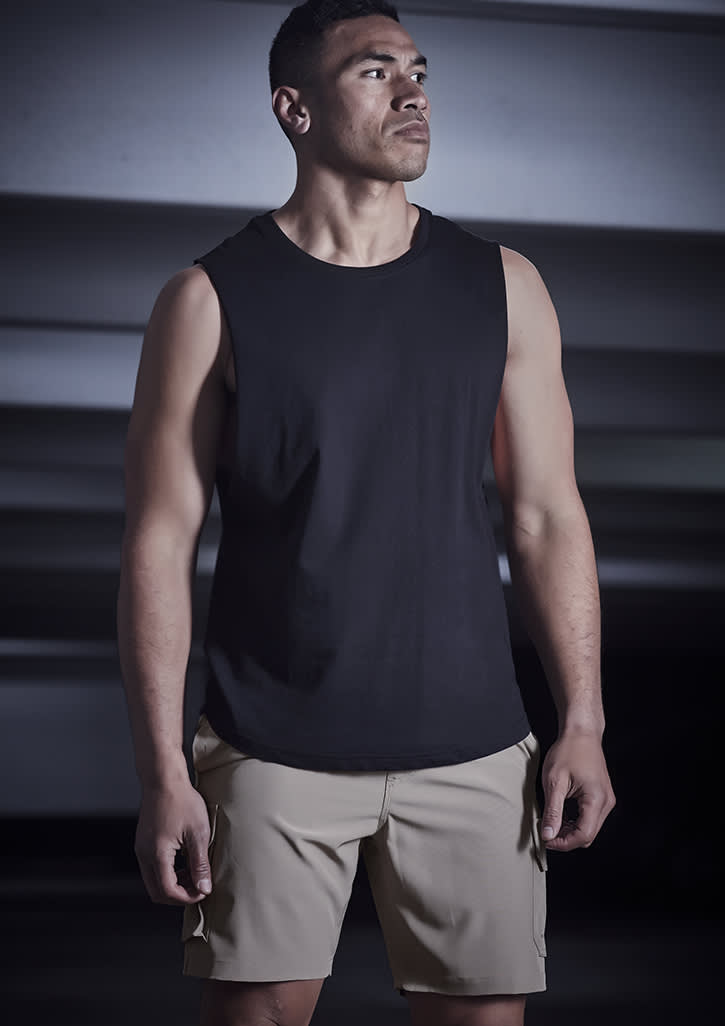 A man wearing a black sleeveless top and beige sports shorts stands in a dimly lit setting. The shorts feature a logo.