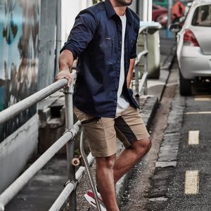 A man in a navy shirt and khaki shorts stands by a railing, with a skateboard and cars in the background.