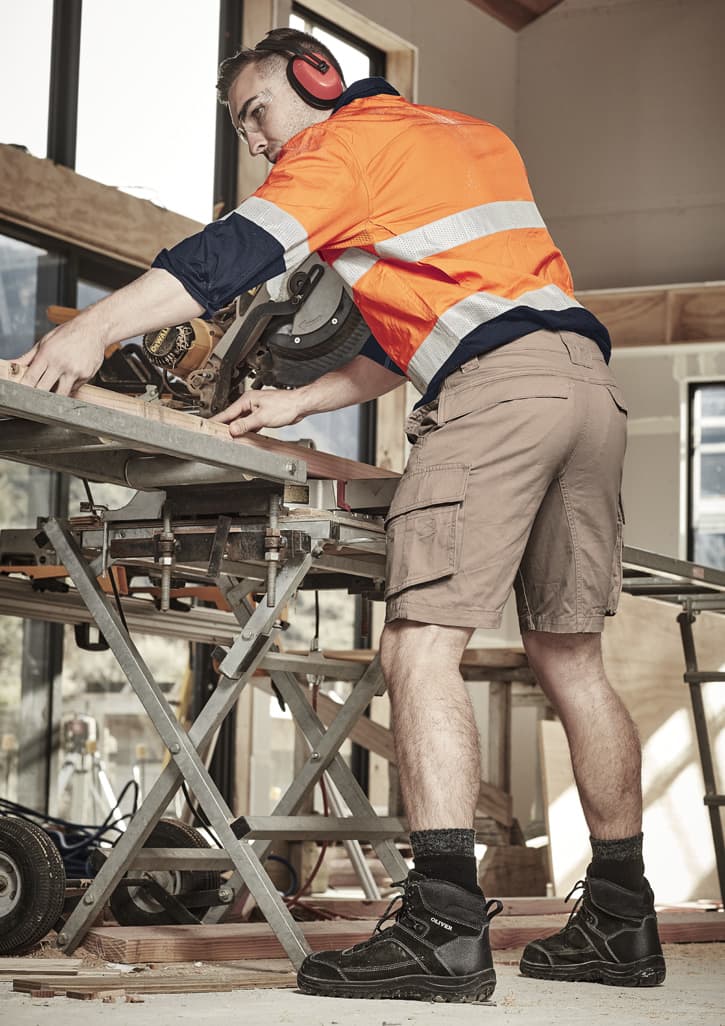 A man wearing beige cargo shorts and a high-visibility orange shirt works with power tools in a construction setting.