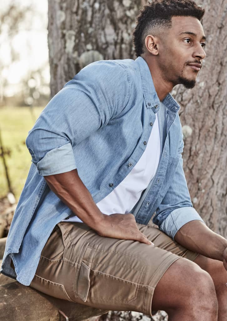A man sits against a tree wearing a light blue button-up shirt and khaki work shorts, showcasing a casual style.
