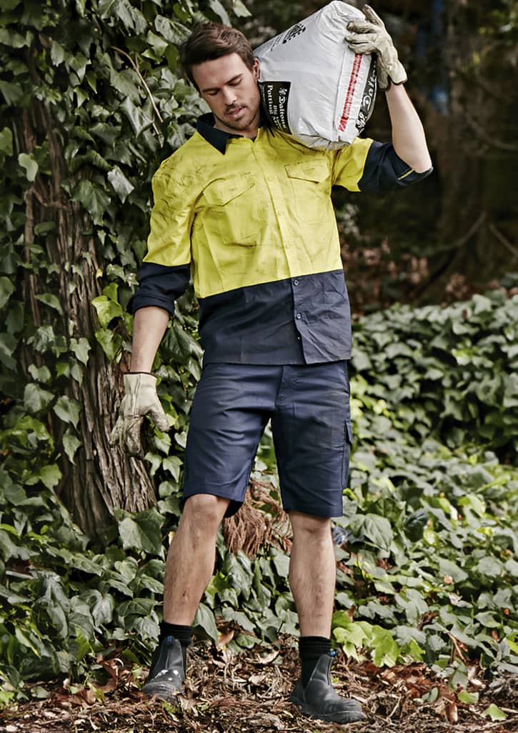 Men's rugged cooling vented shorts in navy, paired with a yellow work shirt, set against a lush green background.