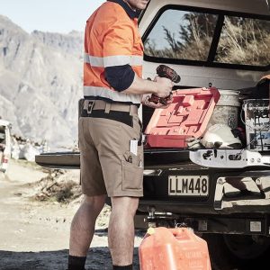 A man wearing orange and navy workwear shorts stands beside a vehicle, holding a drill and a red toolbox.