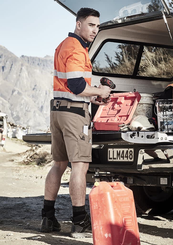 A man wearing orange and navy workwear shorts stands beside a vehicle, holding a drill and a red toolbox.