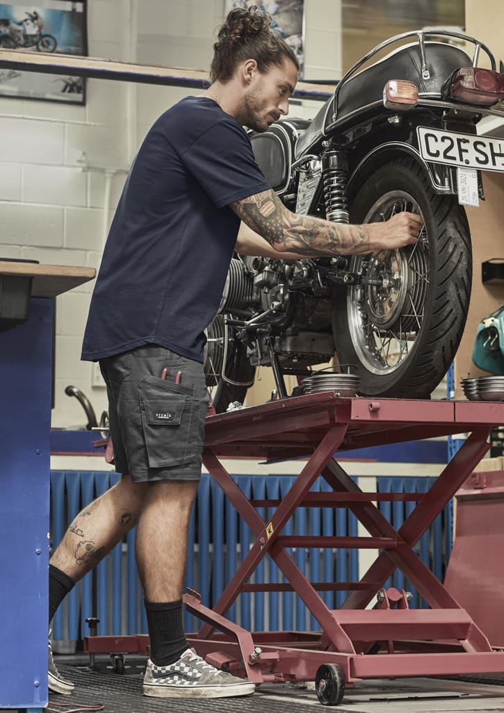 A man in navy shorts works on a motorcycle in a garage, featuring a logo on the shorts.