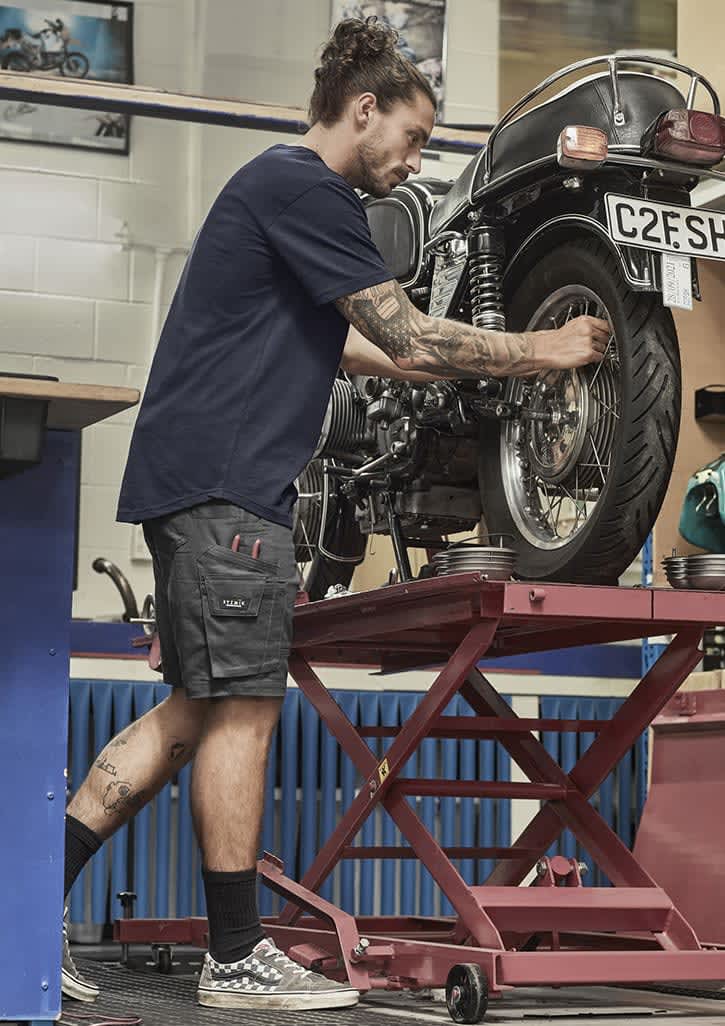 A man in navy blue shorts works on a motorcycle, showcasing rugged cooling stretch cargo shorts with pockets.