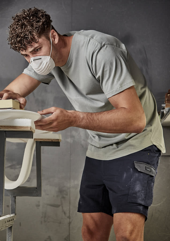 A man wearing a grey t-shirt and navy shorts, working on a project with a mask on in an indoor workspace.
