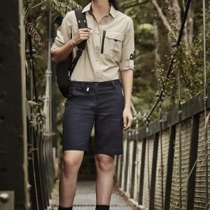 A woman stands on a wooden bridge wearing khaki and navy rugged cooling vented shorts, paired with a button-up shirt and boots.
