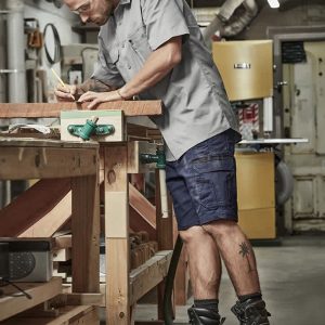 A man wearing grey work shirt and navy shorts stands at a workbench, using a pencil to mark wood. He has ear protection on.