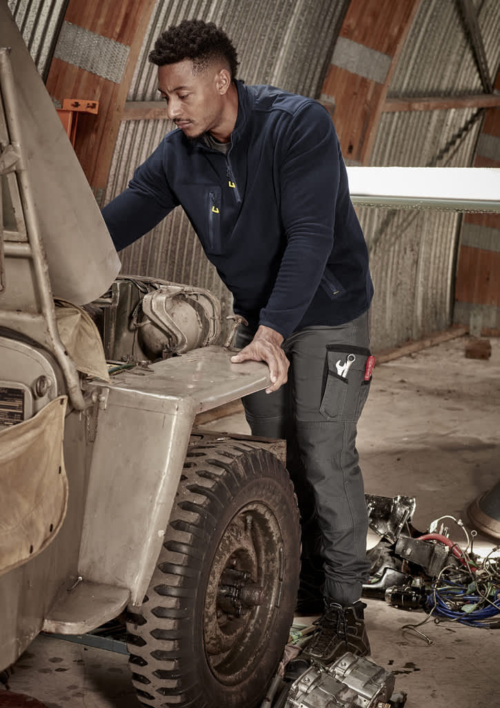 A man in a navy 1/4 zip polar fleece works on an engine in a workshop, wearing work pants with pockets.