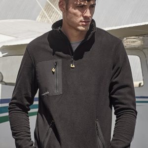 A man wearing a black unisex polar fleece with a quarter-zip and front pockets, set against an aircraft backdrop.