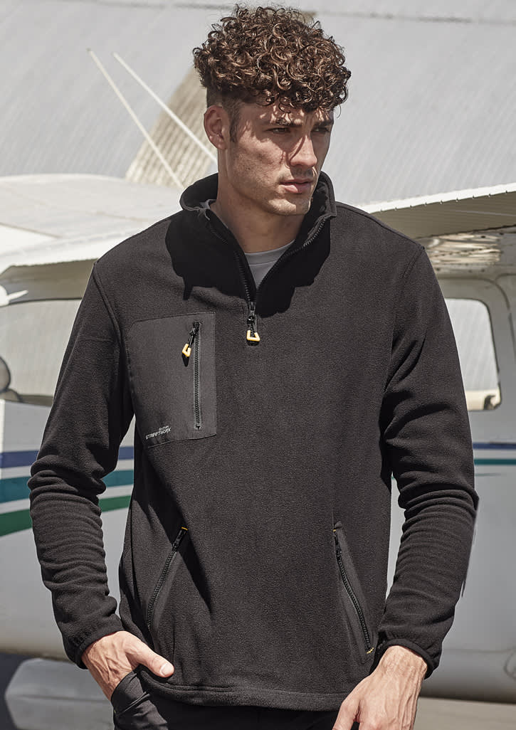 A man wearing a black unisex polar fleece with a quarter-zip and front pockets, set against an aircraft backdrop.