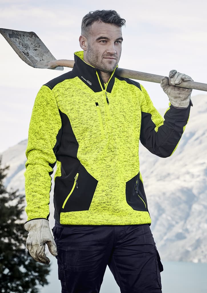 A man holds a shovel while wearing a bright yellow and black reinforced knit 1/2 zip pullover, set against a mountainous backdrop.