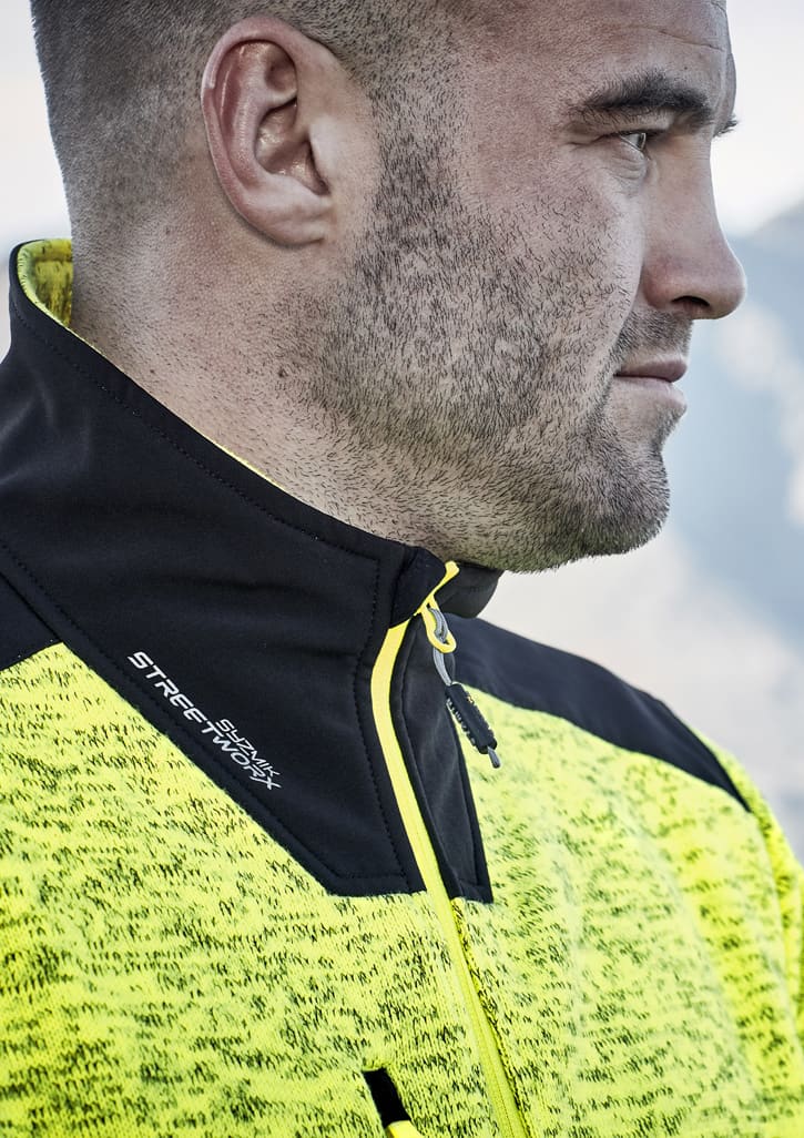 A close-up of a man wearing a bright yellow and black reinforced knit 1/2 zip pullover, featuring a branding logo.