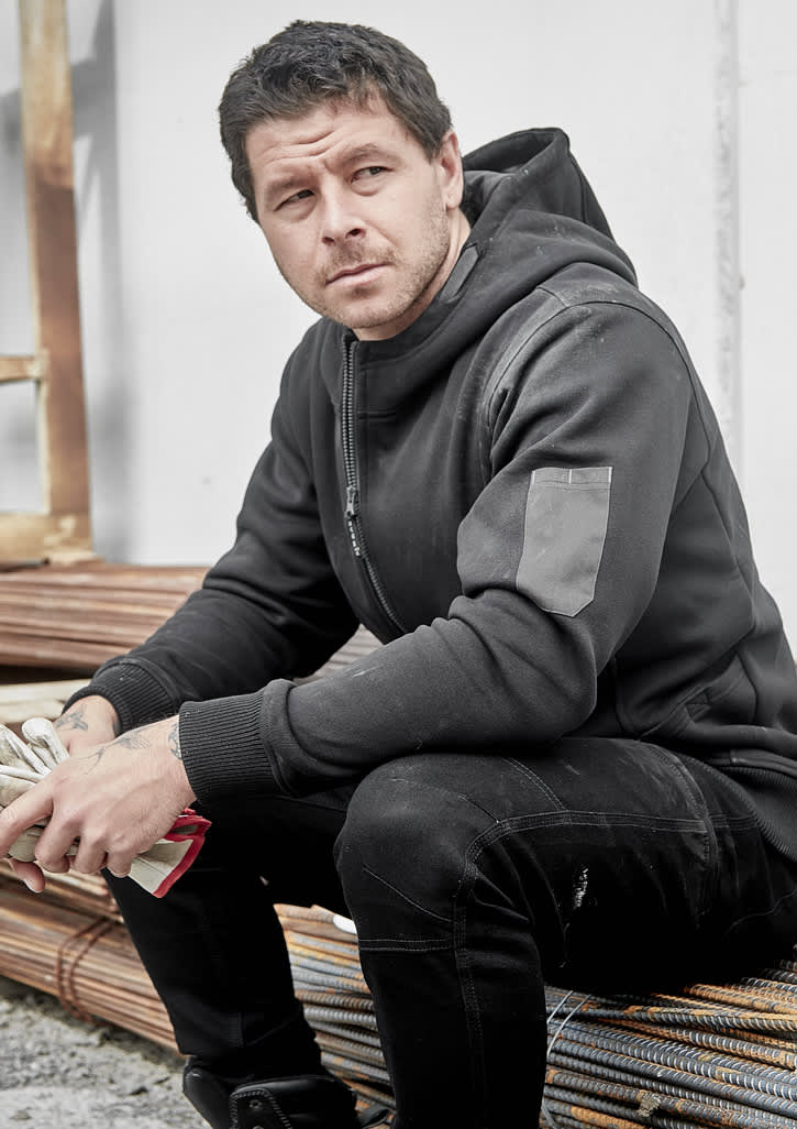 A black unisex zip front hoodie with multi-pockets, featuring a logo, worn by a seated man.
