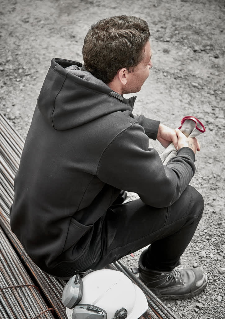 A unisex zip front hoodie in black, featuring multiple pockets, worn by a seated person on a construction site.