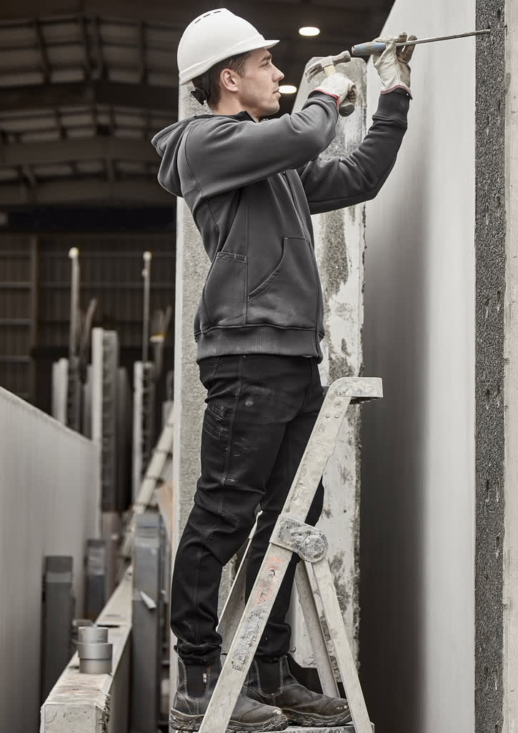 A person in a grey zip front multi-pocket hoodie and black pants using a drill while standing on a ladder.