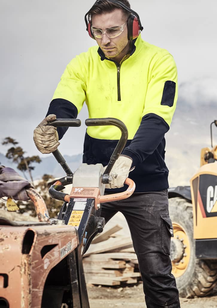A man wears a high visibility yellow and navy unisex 1/2 zip pullover while operating machinery on a work site.