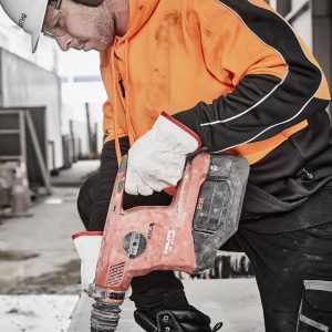 A worker in an orange and black hi-vis pullover hoodie using a power drill while wearing protective gear and gloves.