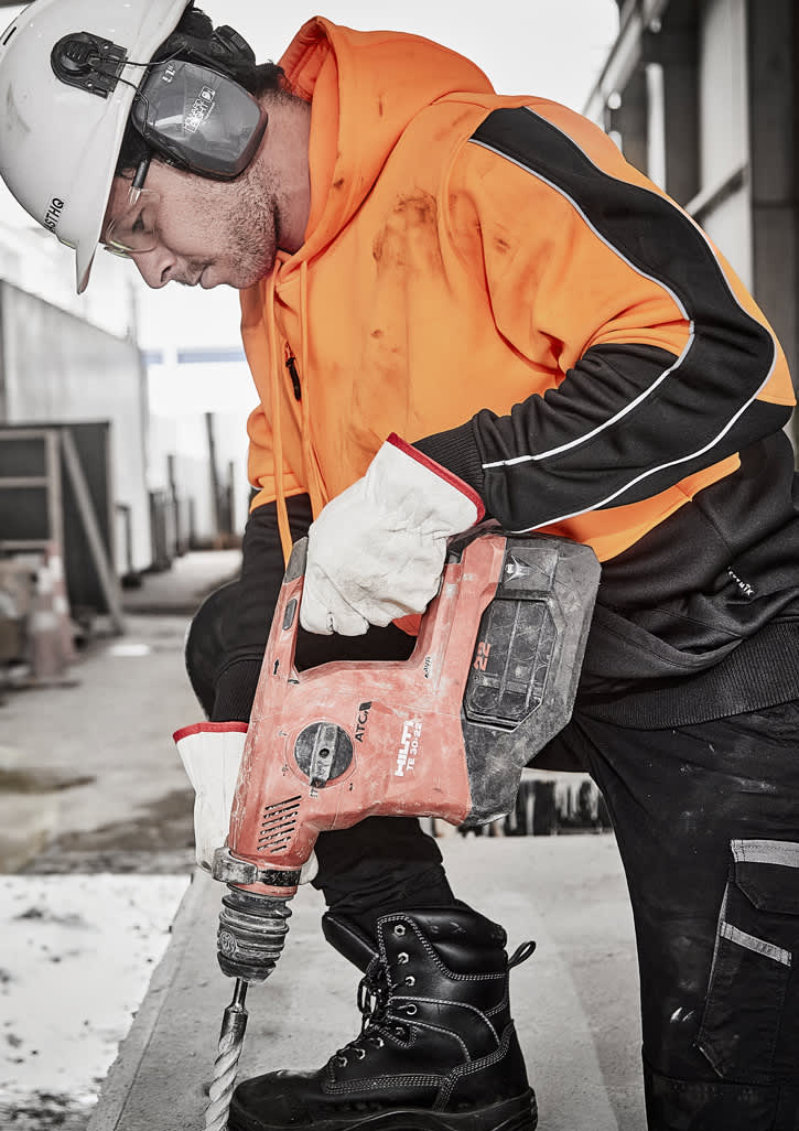 A worker in an orange and black hi-vis pullover hoodie using a power drill while wearing protective gear and gloves.