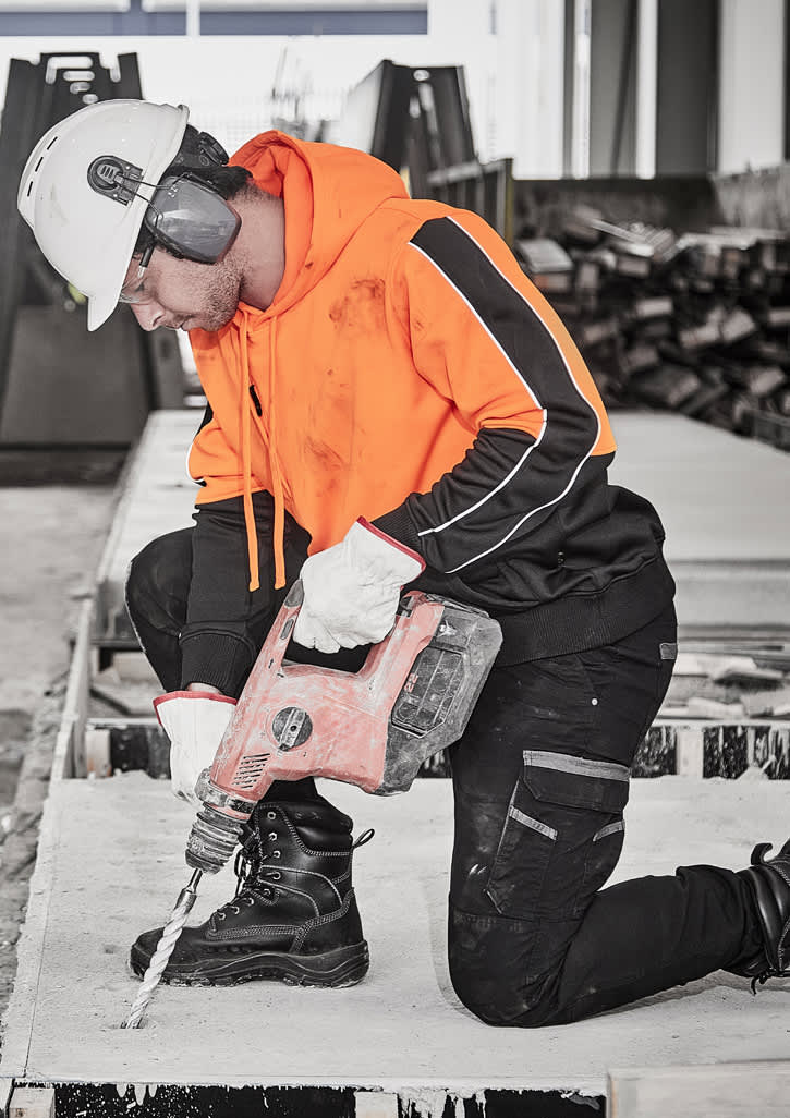 A man in an orange hi-vis pullover hoodie using a drill on a construction site, wearing a safety helmet and ear protection.