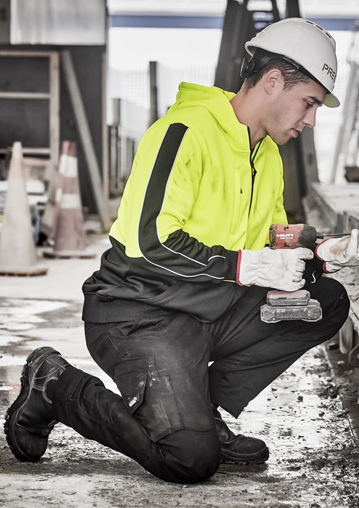 A unisex hi vis full zip hoodie in bright yellow, worn by a person kneeling and working on a construction site.