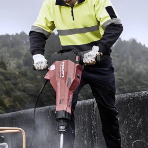A worker in a fluorescent yellow and black hooped pullover operates a demolition tool at a construction site.