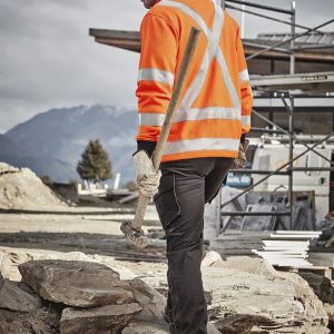 A man wearing an orange high-visibility jumper with reflective stripes, holding a hammer, in a construction setting.