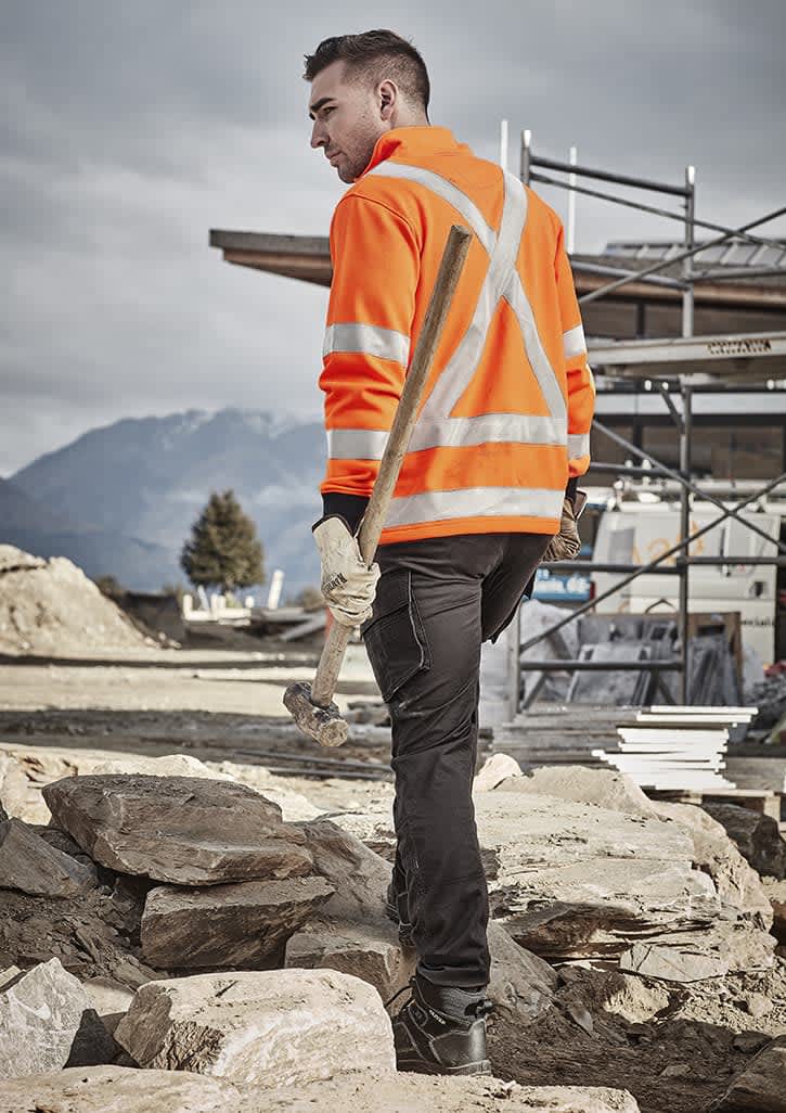 A man wearing an orange high-visibility jumper with reflective stripes, holding a hammer, in a construction setting.