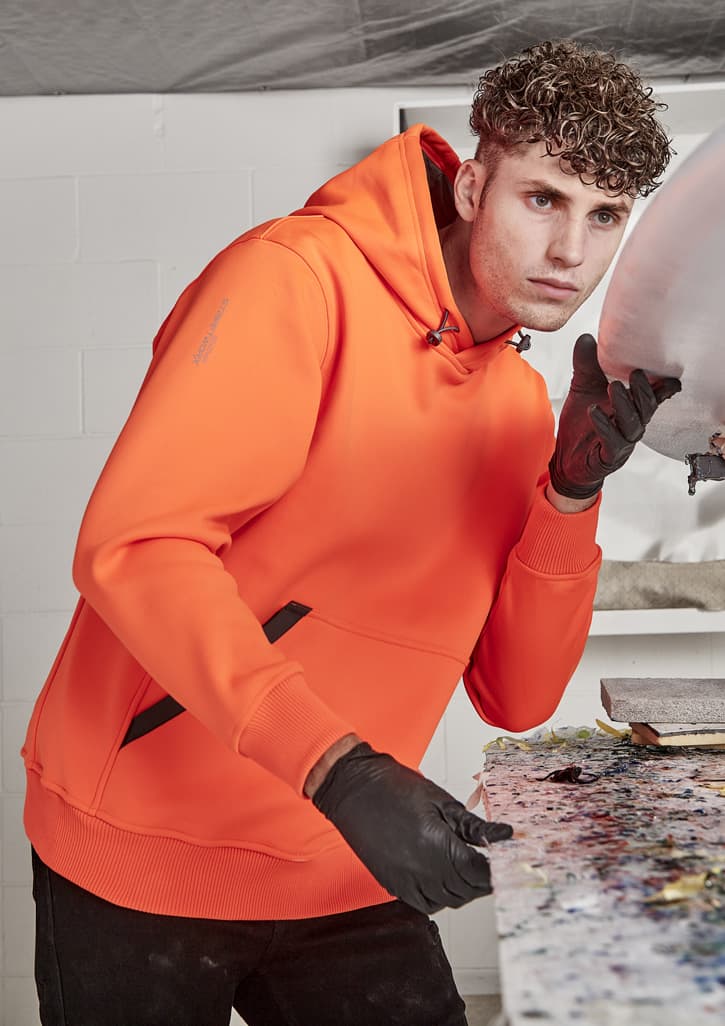 An orange unisex water-resistant hoodie with a black logo, worn by a person in a workshop setting.