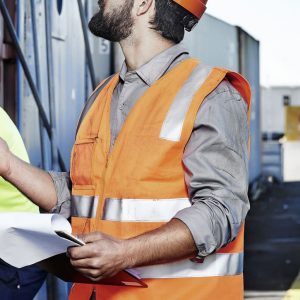 A man wearing an orange hi-vis zip vest with reflective stripes and a hard hat, holding documents at a construction site.