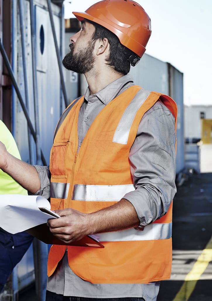 A man wearing an orange hi-vis zip vest with reflective stripes and a hard hat, holding documents at a construction site.