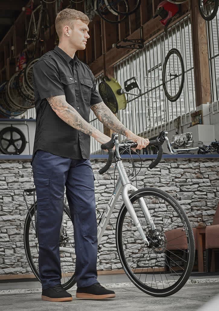 A man in a black shirt stands with a bike, wearing navy midweight drill cargo pants in a workshop setting.