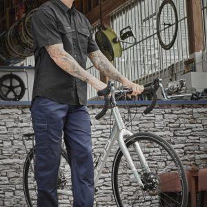 A person wearing navy midweight drill cargo pants and a black shirt stands beside a bicycle in a workshop setting.