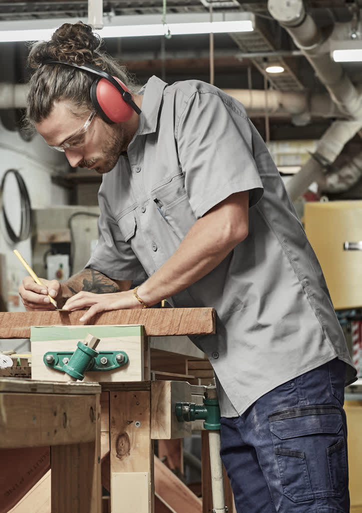 A lightweight grey short sleeve work shirt worn by a man using a pencil on wood in a workshop. The shirt has a logo.