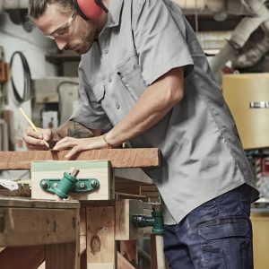 A man wears a grey short sleeve shirt with a logo, using a pencil at a workbench in a workshop setting.