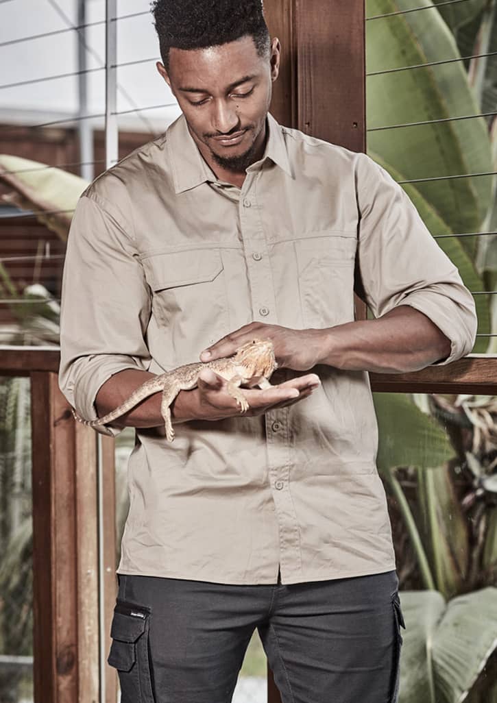 A man wearing a beige long sleeve work shirt holds a lizard in a tropical setting. The shirt features a logo.