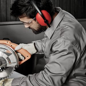 A man in a grey long sleeve work shirt uses a power saw, wearing protective earmuffs and gloves.