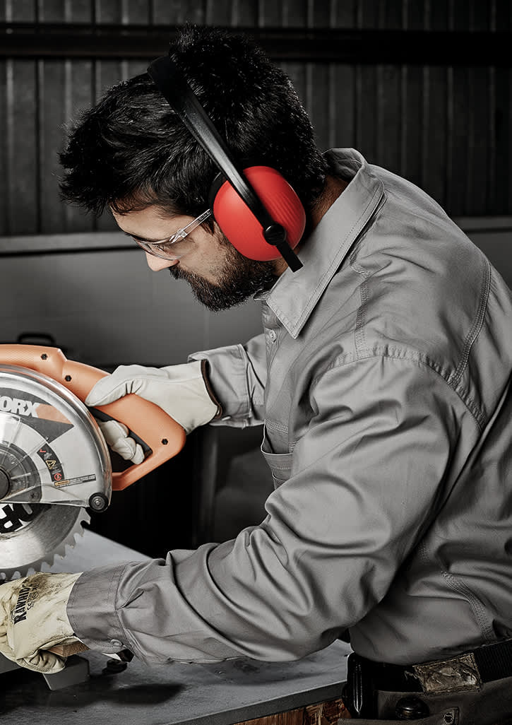 A man in a grey long sleeve work shirt uses a power saw, wearing protective earmuffs and gloves.